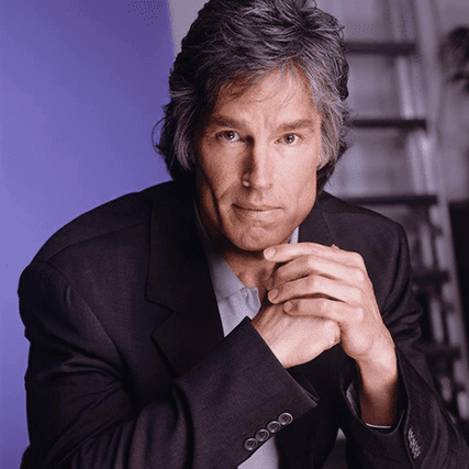 A portrait of a man with graying hair, wearing a dark suit jacket and a light blue shirt, with his hands clasped together, looking directly at the viewer.